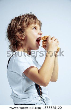 Pretty young curly boy in casual clothes on white studio background. Eating burger. Caucasian male preschooler with bright facial emotions. Childhood, expression, having fun, fast food. Hungry.