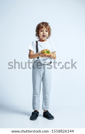 Pretty young curly boy in casual clothes on white studio background. Eating burger. Caucasian male preschooler with bright facial emotions. Childhood, expression, having fun, fast food. Astonished.