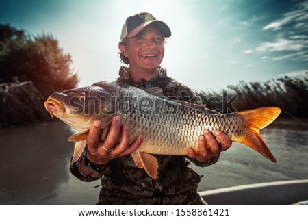 Happy young fisherman holds the big Carp fish (Cyprinus carpio) and smiles Royalty-Free Stock Photo #1558861421