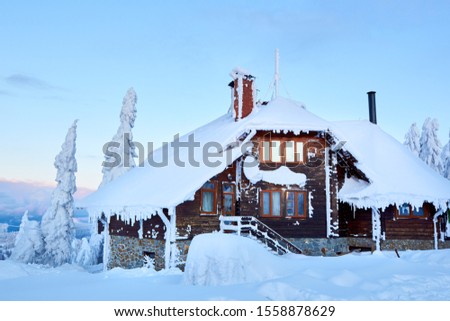 Christmas wooden mansion in mountains on snowfall winter day. Cozy chalet on ski resort near pine forest. Cottage of square timber with wooden facade. Fir-trees covered with snow. Chimney of stone.