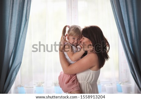 Cute Caucasian mom and daughter, mom holds the girl in her arms and gently hugs kisses, casual style in a real interior