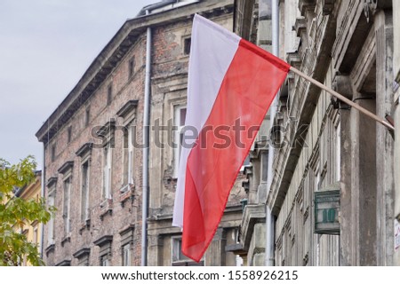 The Polish red and white flag hangs on the facade of an old house on a cloudy day against a gray sky. A national holiday in a country or city, flag day or independence day in Krakow, Poland.
