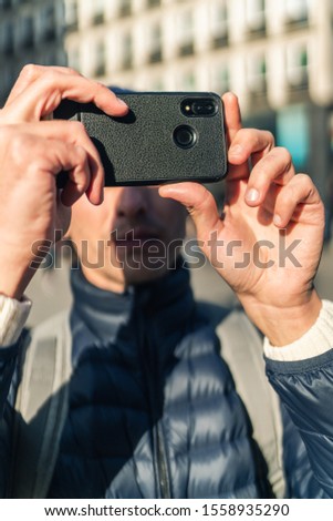 Young man in a cap is taking a photo with his mobile phone on the street of european city.