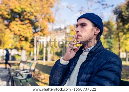 Young man in a cap smoking a cigarette on a bench during a nice autumn day.