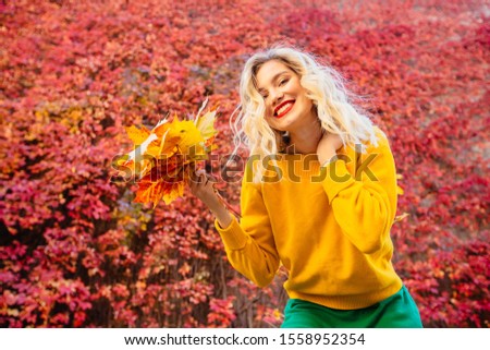 Emotional portrait of cheerful playful young blond caucasian woman in yellow sweater, red lips, waving at camera autumn leafs in front of red foliage on background. Autumn mood concept.