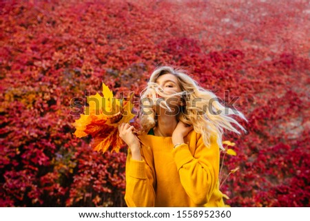 Emotional portrait of cheerful playful young blond caucasian woman in yellow sweater, red lips, waving at camera autumn leafs in front of red foliage on background. Autumn mood concept.