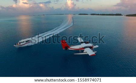 Aerial photo of seaplane during sunset. White black red seaplane docked in the middle of the Maldivian lagoon of Indian Ocean, far away from Island. Speed boat is passing by