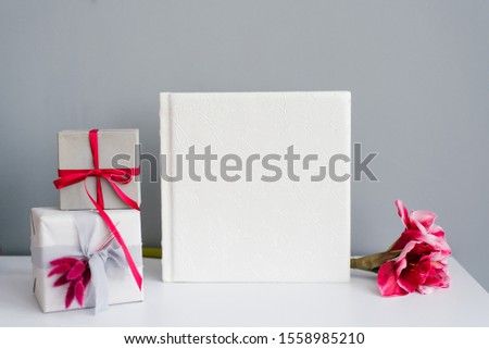 Classic photo album in a leather white cover surrounded by gift boxes and a pink flower on a gray background