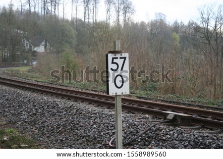 What can be seen, is the picture of train-tracks with a sign by the side. The weather is grey and it seems kind of cold.