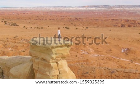 AERIAL: Young woman traveler stands atop a cliff and observes the stunning canyon in United States. Female photographer looking for inspiration while observing desert nature from a massive boulder.