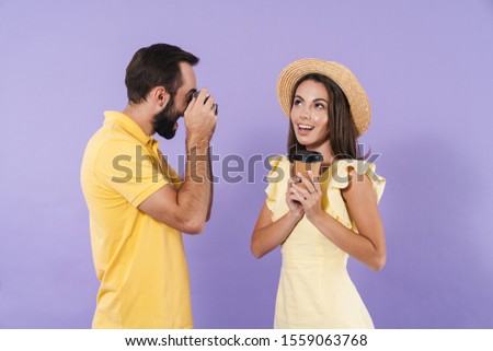 Happy excited beautiful young couple wearing casual clothing standing isolated over violet background, man taking photo of his girlfriend