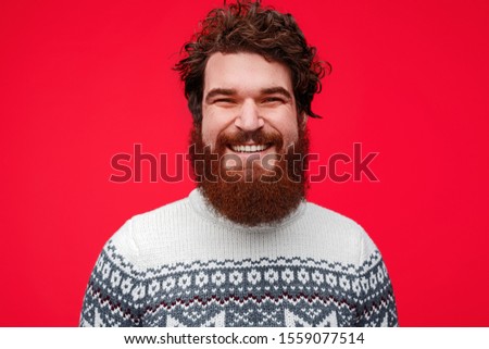 Delighted bearded male in warm knitted sweater smiling and looking at camera while standing against red background on winter day