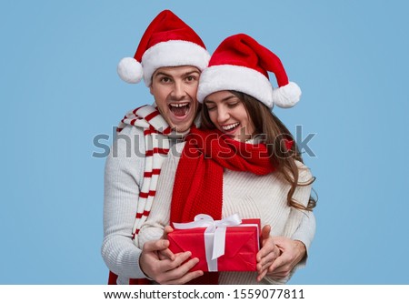 Delighted and surprised young man and woman in Santa hat and warm clothes examining wrapped gift box during Christmas party