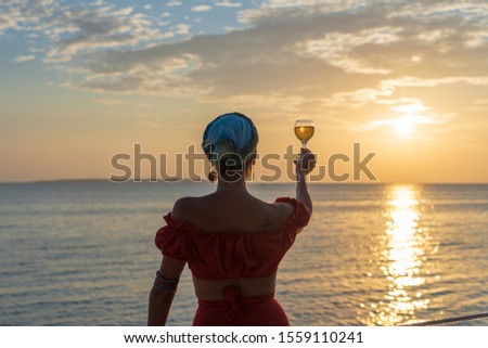 Cute woman holding glass of white wine against a beautiful sunset near sea waves on the tropical beach, close up. Girl is enjoying the sunset with a glass of wine. Concept of leisure and travel