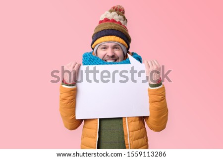Smiling young man in warm winter hats holding blank sign in hands on pink background.