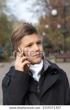 Portrait of a schoolboy dressed in a business suit talking on a cell phone, holding it in his hand near his ear. Front view with a serious expression and a slight smile. Autumn day in the boulevard