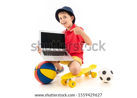 Caucasian teenager boy sits on a yellow penny with basketball and soccer balls and shows his laptop