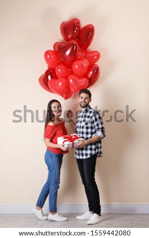 Happy young couple with gift box and heart shaped balloons near beige wall. Valentine's day celebration