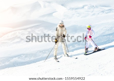 Young adult beautiful sporty mother having fun skiing with kid daughter on mountain alpine ski winter resort. Slim mom in luxury fashion skier suit and child enjoy outdoor recreation activities