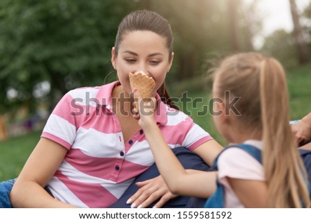 Close up image of young happy family spending their weekend in park, relaxing on frameless chair, charming daughter feeding her moomy with ice cream, enjoing to spend time together, erxpressing joy.