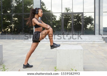 Side view of dark skinned sporty female in black athletic clothes and sneakers listening to music with headphones while doing physical exercises outdoor. Concept for sports and healthy lifestyle