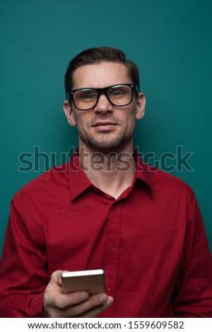 Close-up - Portrait of a positive young man in glasses and a red shirt holding a smartphone in his hands on a green background. Advertising concept and place for text
