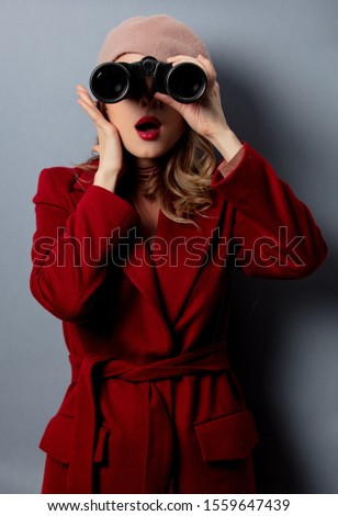 Young woman in red coat with binoculars on grey background 