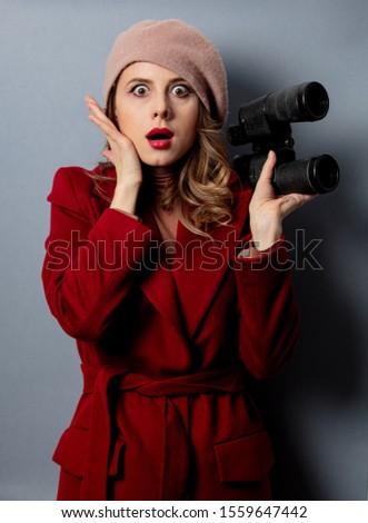 Young woman in red coat with binoculars on grey background 