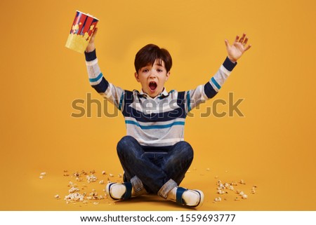 Cute little boy sitting and watching movie and eats popcorn against yellow wall.