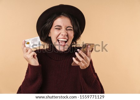 Image of screaming emotional young brunette woman posing isolated over beige wall background holding credit card using mobile phone.