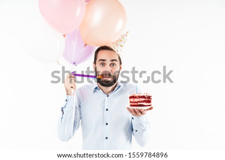 Image of young man blowing party horn and holding birthday cake with air balloons isolated over white background