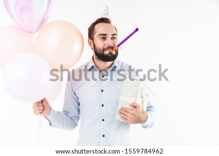 Image of beautiful man blowing party horn while holding air balloons and present box isolated over white background