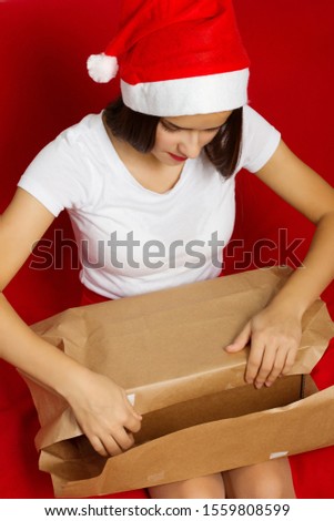 Christmas mood. A young European girl with long dark hair in a Santa Claus hat is sitting on the sofa and packages new year gifts