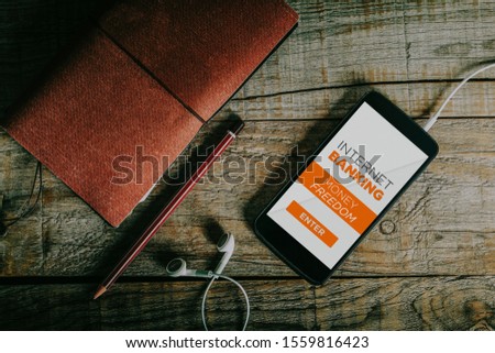 Top view of cellphone with internet banking service app in the screen. Mobile on a wooden desk, notebook, pencil and earphones.