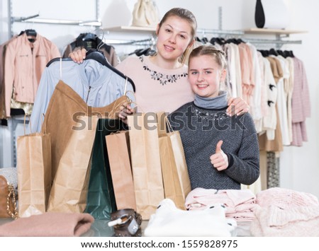 Excited mother and daughter holding shopping bags and showing thumbs up in clothes shop
