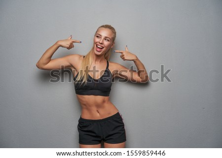 Cheerful attractive young sporty blonde woman with casual hairstyle pointing to herself with forefingers and looking happily aside with wide smile, isolated over light grey background