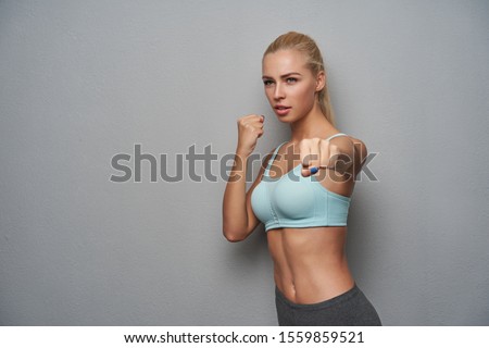 Severe sporty young blonde woman with casual hairstyle looking aside and boxing with raised fists, having hard work out after working day, standing against light grey background