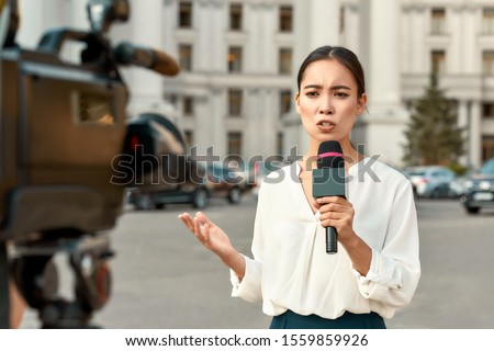 Cropped portrait of professional female reporter at work. Young woman standing on the street with a microphone in hand and smiling at camera. Horizontal shot. Selective focus on woman Royalty-Free Stock Photo #1559859926