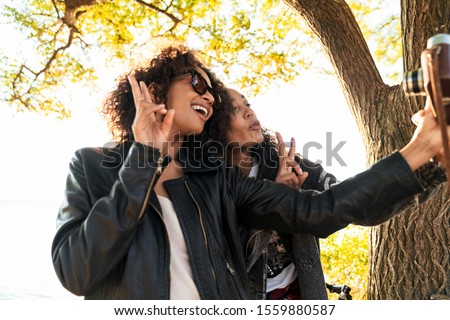 Image of two joyful african american girls in trendy streetwear smiling and taking selfie on retro camera outdoors