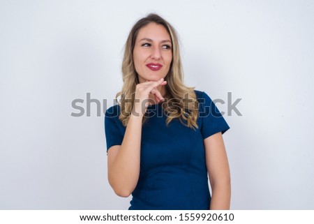 Dreamy  female with pleasant expression, wearing casual clothes, looks sideways, keeps hand under chin, thinks about something pleasant, poses against gray background.