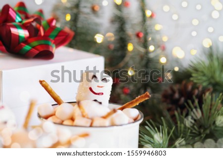 Two snowmen soaking in a hot cup of cocoa surrounded by mini marshmallows. Extreme selective focus on snowman's face with blurred foreground and background. Christmas trees and gift in background.