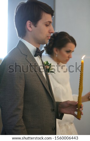 Bride and groom standing at wedding ceremony. Happy stylish wedding couple holding candles with light under golden crowns during holy matrimony in church.