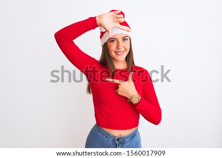 Young beautiful girl wearing Christmas Santa hat standing over isolated white background smiling making frame with hands and fingers with happy face. Creativity and photography concept.