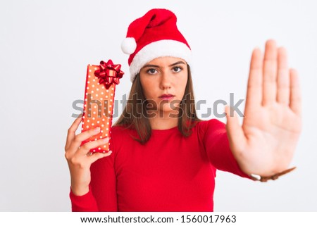 Young beautiful girl wearing Christmas Santa hat holding gift over isolated white background with open hand doing stop sign with serious and confident expression, defense gesture