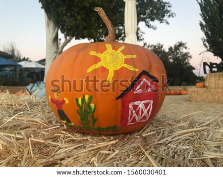Orange pumpkin on hay stack with colorful illustration of a farm house, corn stalk, scarecrow, and yellow sun.