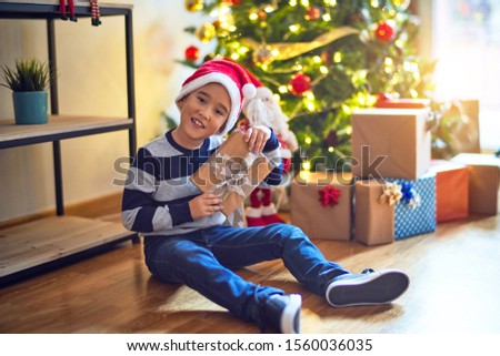 Adorable toddler smiling happy and confident. Sitting on the floor wearing santa claus hat with smile on face holding gift around christmas tree at home