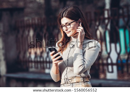 Image of beautiful stylish woman holding mobile phone. Young european girl standing at the street and using cellphone. Woman listening to music with the phone and having fun. Mobile internet concept. 