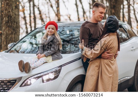 Happy family in the autumn forest. Daughter sits on the hood of a new car, dad and mother hug and kiss. 