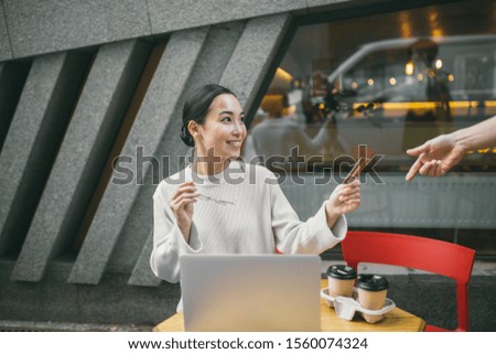 Young handsome Asian woman with glasses in hand is sitting in a cafe outside and giving her credit card to a waiter.