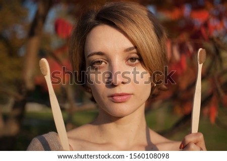 Young girl with short blond hair is holding a bamboo toothbrushes outdoors during sunny weather in autumn on red and orange leaves background. Environmental friendliness and zero waste concept